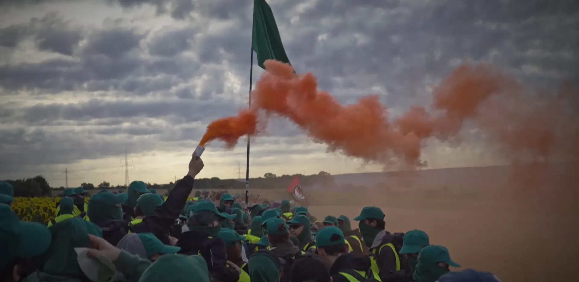 Grüner Finger. Menschen mit grünen Kappis und Schlauchschals. Eine Person hält einen roten Rauchtopf. Im Hintergrund sind weit entfernt Felder zu sehen. Eine Person schwenkt eine Antifa-Fahne