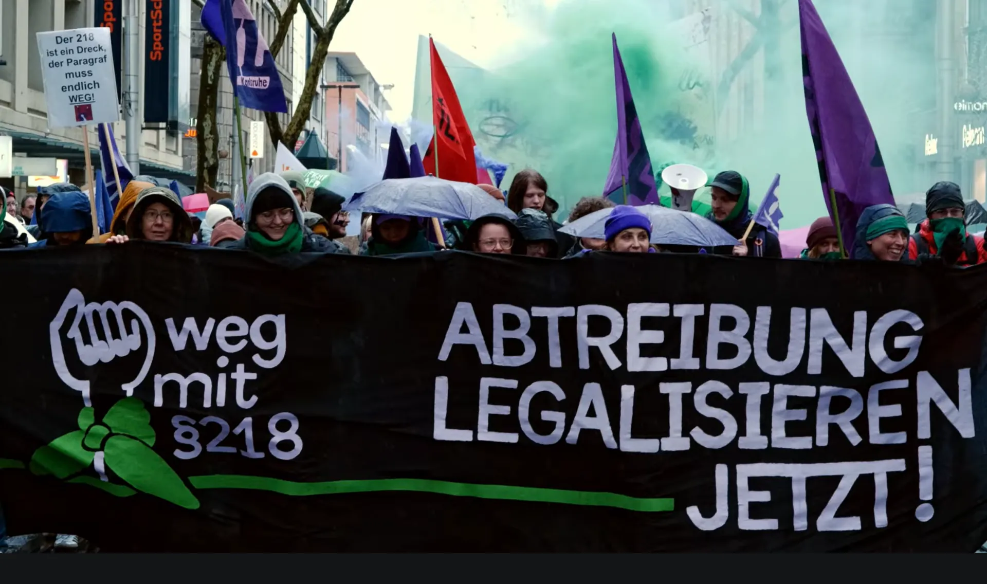 Demonstration in Karlruhe. Banner mit Faust und Abtreibungen Legalisieren Jetzt. Dahinter Menschen, Fahnen und grüner Rauch