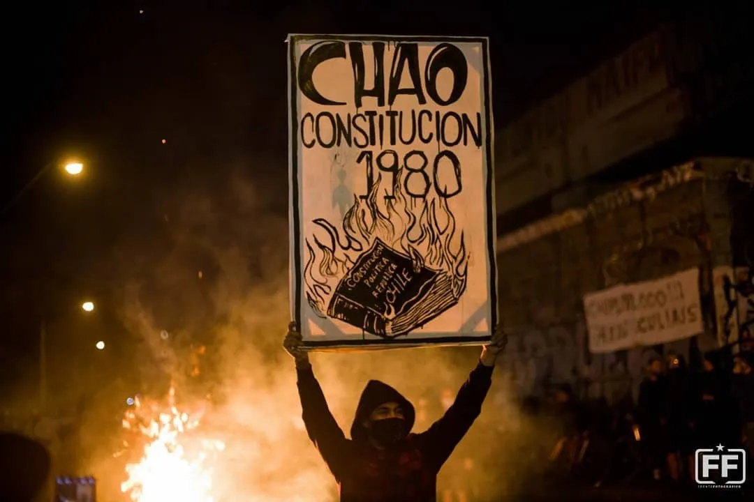 ein vermummter Demonstrant hält ein Schild mit der Aufschrift Chao Constitucion 1980, im Hintergrund Feuer und Rauch
