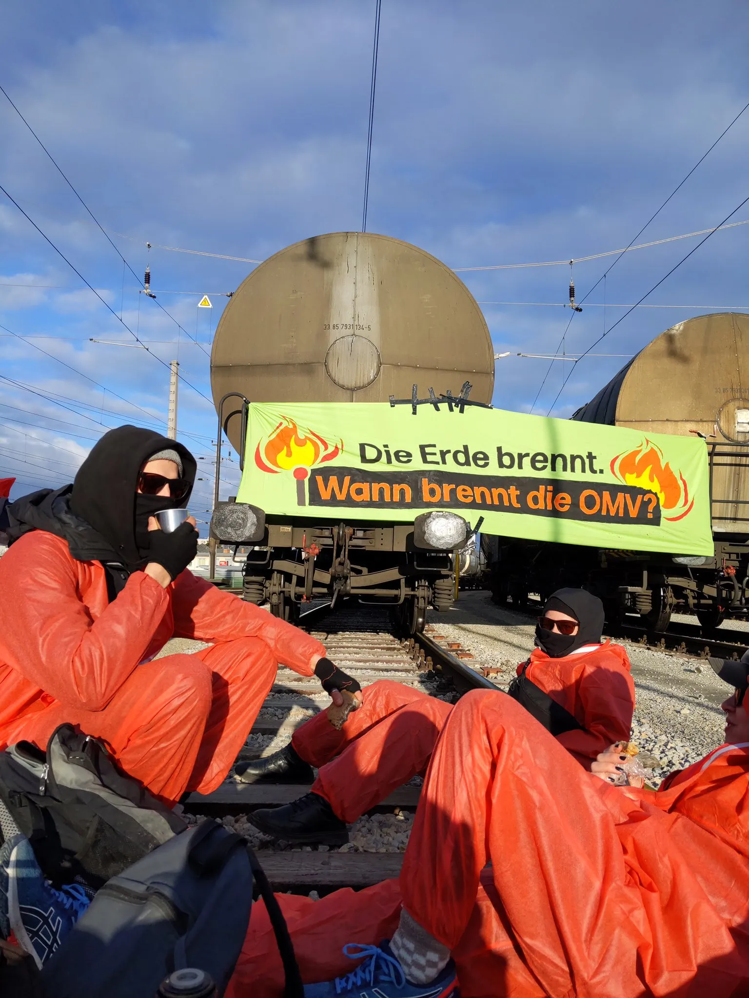 Menschen auf Schienen in orangenen Anzügen vor einem Banner mit der Aufschrift Die Erde brennt wann brennt die OMV?