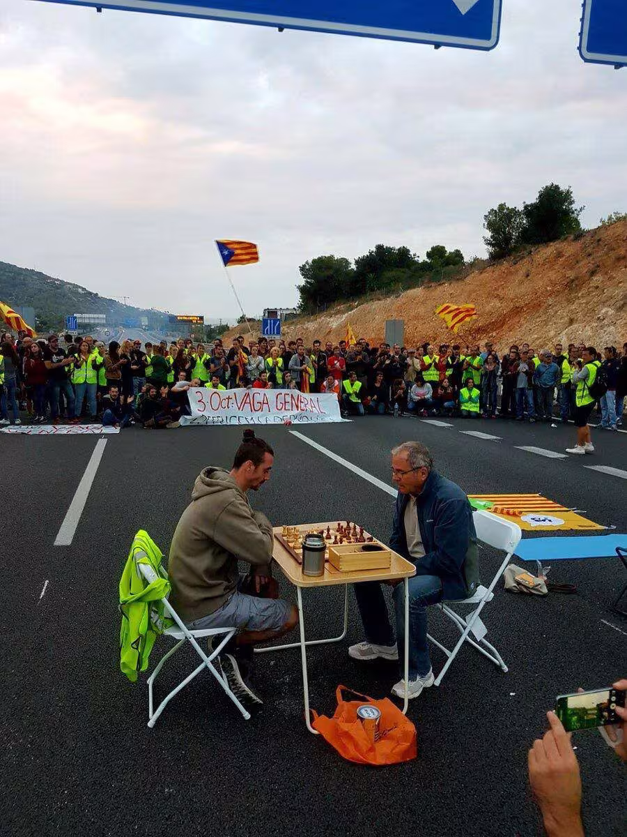 Straßenblockade auf Autobahnen. Menschen schwenken katalanische Flaggen. Zwei Menschen stizen auf Stühlen an einem Tisch und spielen Schach