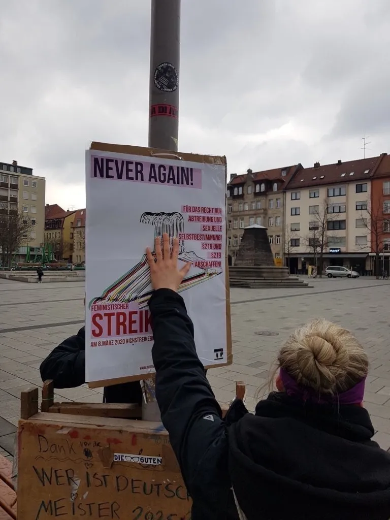 Person plakatiert IL Plakat an Laterne "Never Again - Feministischer Streik"