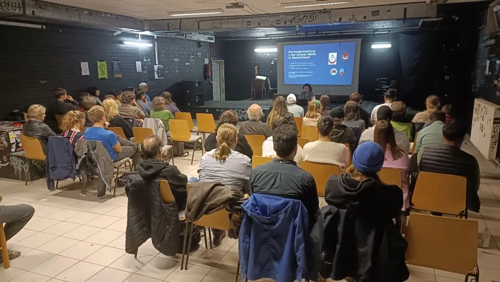 Foto eines Vortrags. Menschen sitzen auf Stühlen und schauen auf einen Beamer