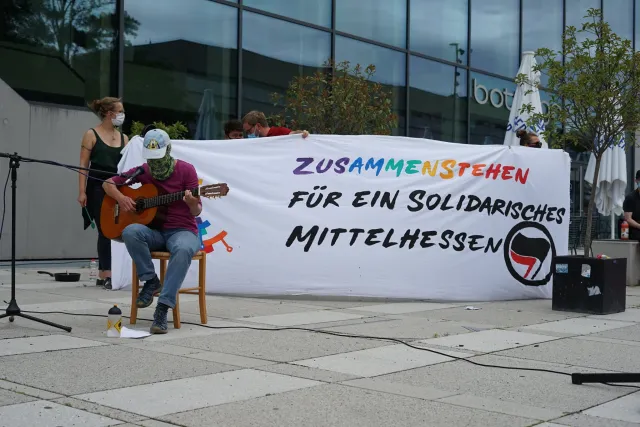 Person spielt vermummt gitarre mit mikrofon dahinter banner: zusammen für ein solidarisches mittelhessen