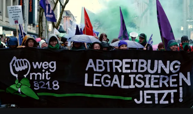 Demonstration in Karlruhe. Banner mit Faust und Abtreibungen Legalisieren Jetzt. Dahinter Menschen, Fahnen und grüner Rauch