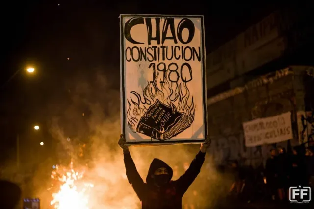 ein vermummter Demonstrant hält ein Schild mit der Aufschrift Chao Constitucion 1980, im Hintergrund Feuer und Rauch
