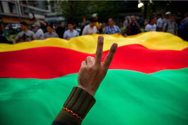 eine Hand zum Peace-Zeichen geformt, dahinter eine Rojava Fahne