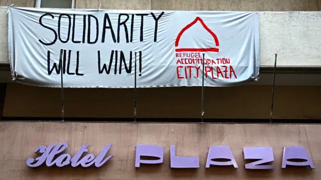 Banner hängt vor einem Haus mit der Aufschrift "Solidarity will win", daneben "Refugee Accomodation City Plaza"