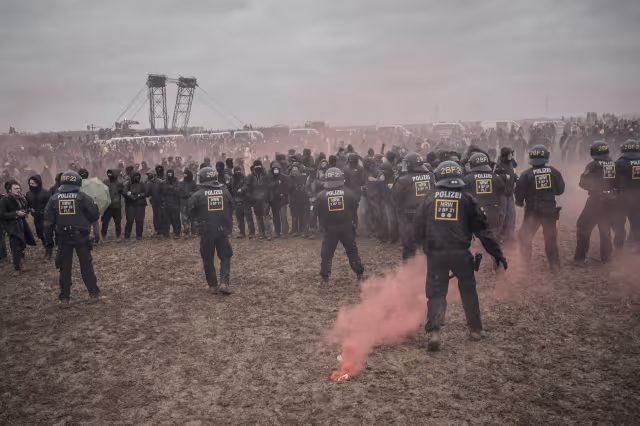 auf einer schlammigen Fläche stehen sich eine Kette von schwarz gekleideten, teilweise vermummten Demonstrierenden und Cops gegensüber, im Vordergrund brennt rosa Pyro