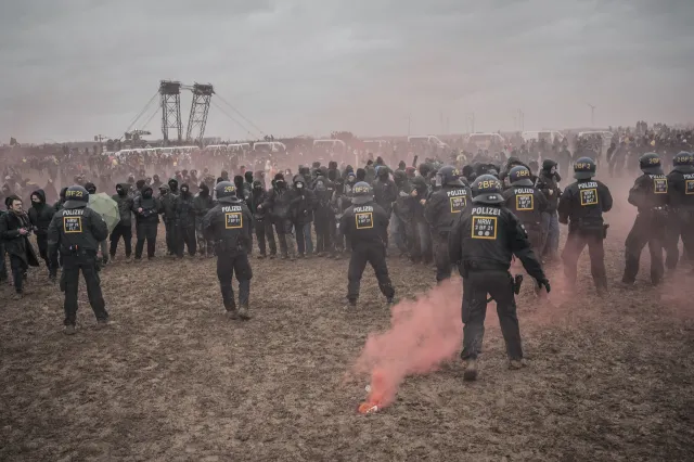auf einer schlammigen Fläche stehen sich eine Kette von schwarz gekleideten, teilweise vermummten Demonstrierenden und Cops gegensüber, im Vordergrund brennt rosa Pyro