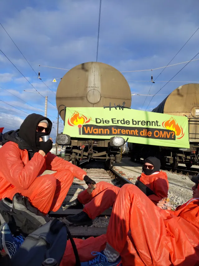 Menschen auf Schienen in orangenen Anzügen vor einem Banner mit der Aufschrift Die Erde brennt wann brennt die OMV?