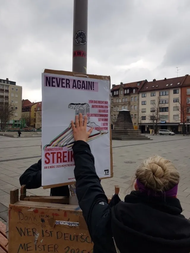 Person plakatiert IL Plakat an Laterne "Never Again - Feministischer Streik"