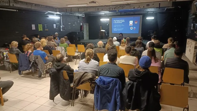 Foto eines Vortrags. Menschen sitzen auf Stühlen und schauen auf einen Beamer